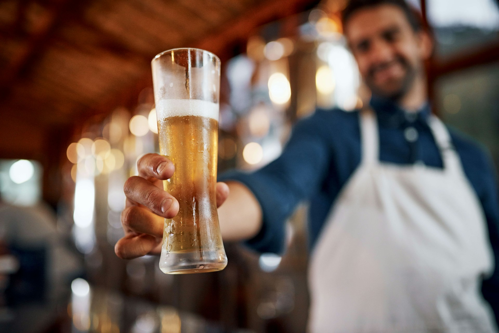 Closeup of a cheerful young business owner holding up a glass of beer that he just poured, taken inside a brewery during the day